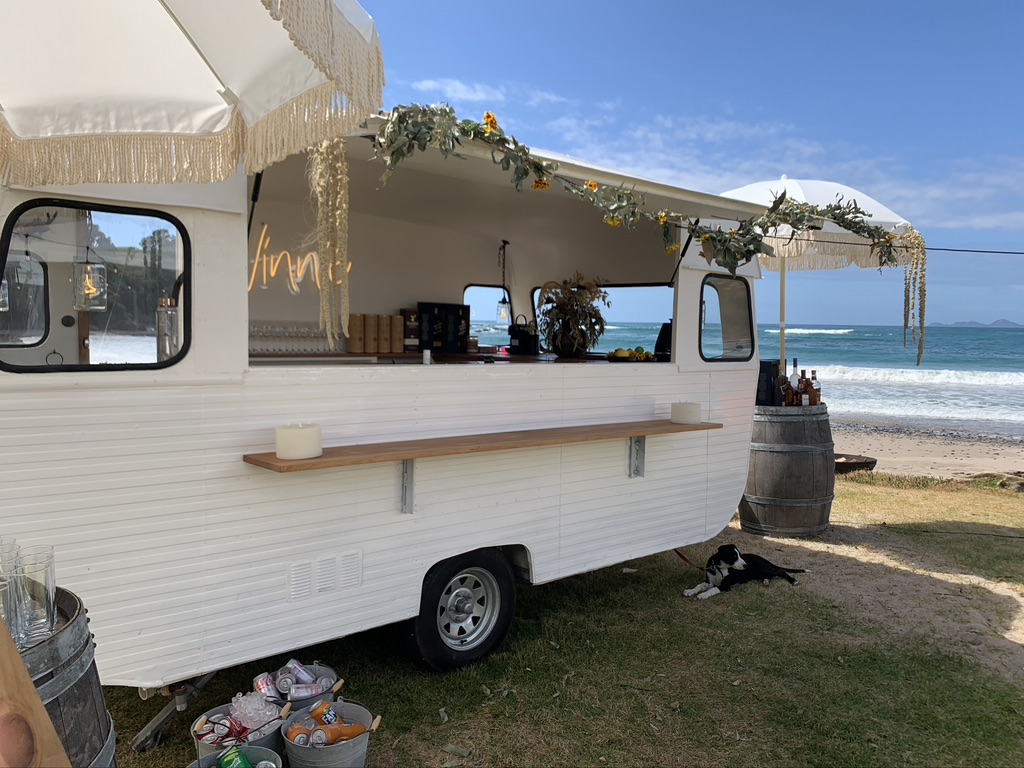 Vinnie the vintage caravan bar set up on the beach with a fringed awning and floral garland